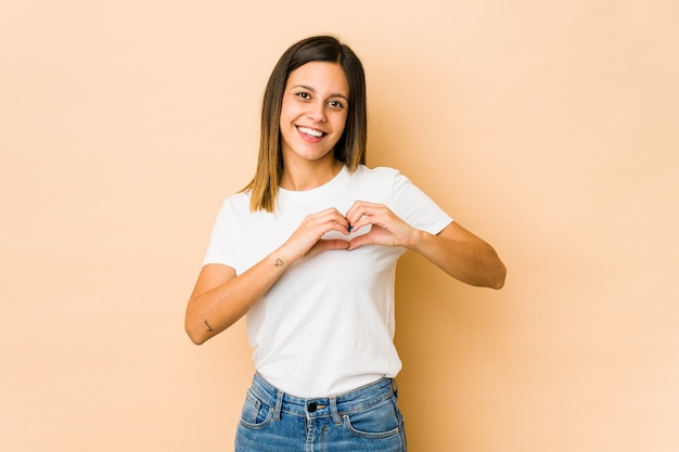 Jeune femme isolée sur beige souriant et montrant une forme de coeur avec les mains.