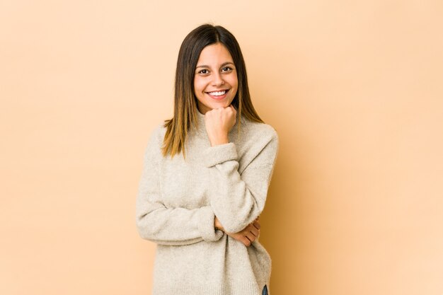 Jeune femme isolée sur fond beige souriant heureux et confiant, touchant le menton avec la main.