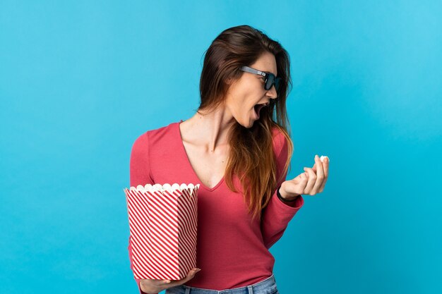 Jeune femme isolée sur un mur bleu avec des lunettes 3D et tenant un grand seau de pop-corn tout en regardant côté