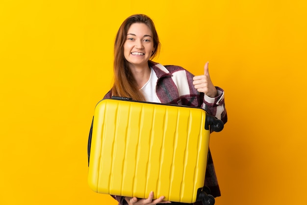 Jeune femme isolée sur un mur jaune en vacances avec valise de voyage et avec le pouce vers le haut