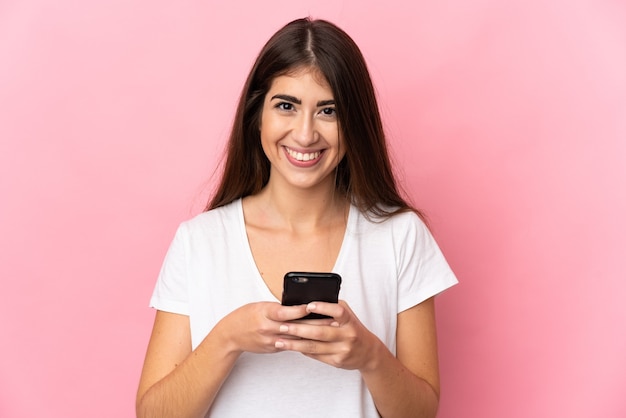 Jeune femme isolée sur un mur rose en regardant la caméra et souriant tout en utilisant le mobile