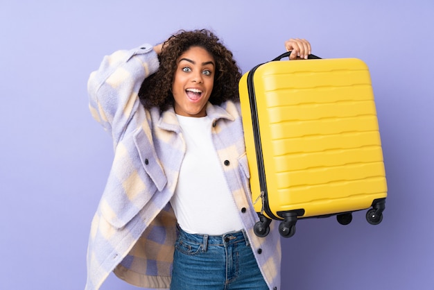 Jeune femme isolée sur un mur violet en vacances avec valise de voyage et surpris