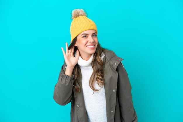 Jeune femme italienne portant une veste d'hiver et un chapeau isolé sur fond bleu écoutant quelque chose en mettant la main sur l'oreille