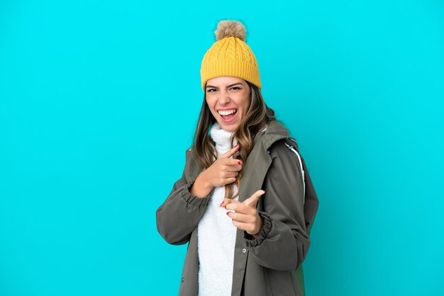 Jeune femme italienne portant une veste d'hiver et un chapeau isolé sur fond bleu pointant vers l'avant et souriant