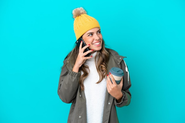 Jeune femme italienne portant une veste d'hiver et un chapeau isolé sur fond bleu tenant du café à emporter et un mobile