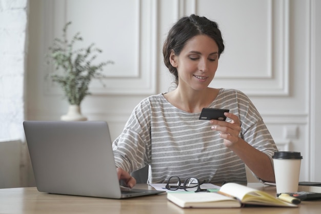 Jeune femme italienne souriante dans des vêtements décontractés regardant une carte de crédit et tapant sur un ordinateur portable