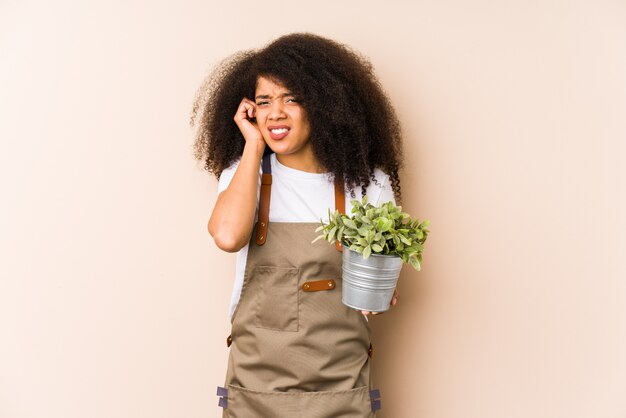 Jeune femme jardinier afro tenant une plante isolée couvrant les oreilles avec les mains.