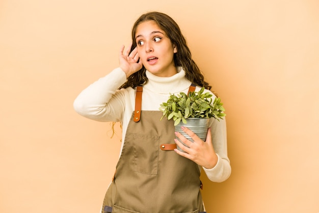 Jeune femme de jardinier caucasien tenant une plante isolée en essayant d'écouter un commérage.