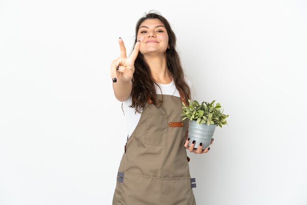 Jeune femme jardinier russe tenant une plante isolée souriant et montrant le signe de la victoire