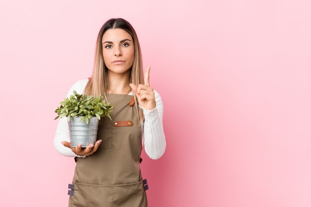 Jeune femme jardinier tenant une plante montrant le numéro un avec le doigt.