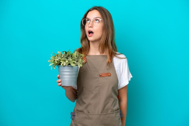 Jeune femme jardinière caucasienne tenant une plante isolée sur fond bleu en levant et avec une expression surprise