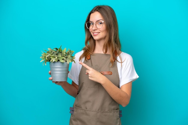 Jeune femme jardinière caucasienne tenant une plante isolée sur fond bleu pointant vers le côté pour présenter un produit