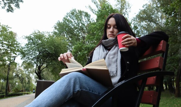 Jeune Femme En Jeans, Manteau Et écharpe, Sur Un Banc De Parc. Une Femme Lit Un Livre Et Boit Du Café Ou Une Autre Boisson Chaude Seule à L'extérieur.