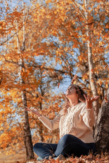 Jeune femme jetant des feuilles jaunes en l'air assis dans la forêt d'automne