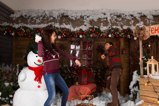 Jeune femme jetant de la neige de coton à son partenaire tout en jouant à l'intérieur de la maison en bois avec un bonhomme de neige et un assortiment de belles décorations de Noël.