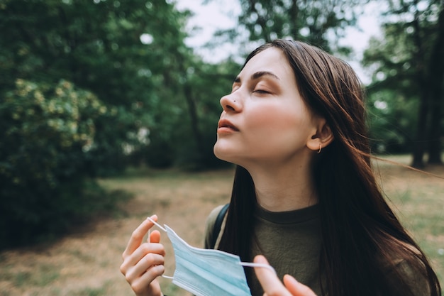 Jeune femme jolie caucasienne enlève un masque de protection médicale de son visage sur la nature, respirant de l'air frais et pur.