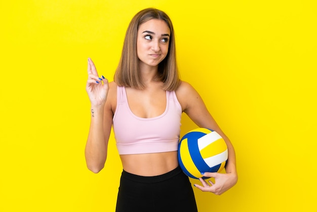 Jeune femme jouant au volley-ball isolé sur fond jaune avec les doigts croisés et souhaitant le meilleur