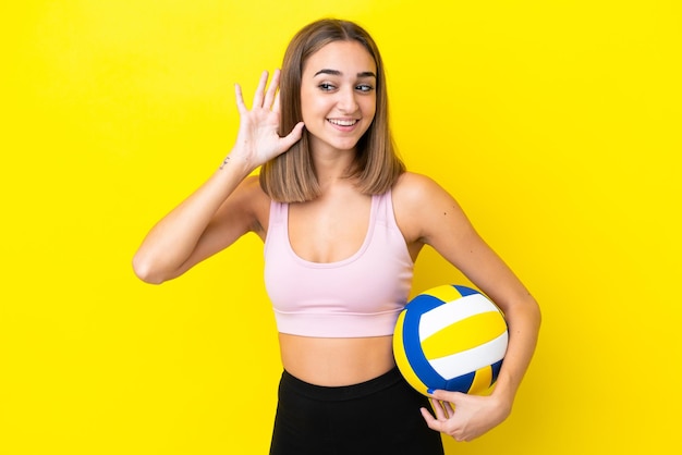 Jeune femme jouant au volley-ball isolée sur fond jaune écoutant quelque chose en mettant la main sur l'oreille