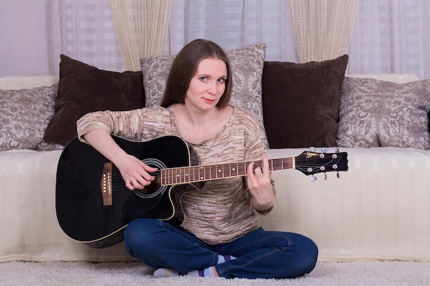 Une jeune femme joue d'une guitare acoustique noire sur le tapis de la chambre