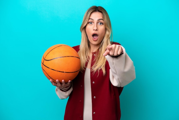 Jeune femme de joueur de basket-ball caucasien isolé sur fond bleu surpris et pointant vers l'avant