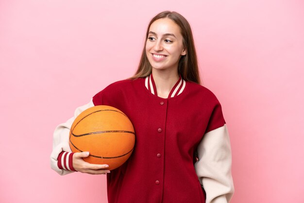 Jeune femme de joueur de basket-ball caucasien sur isolé sur fond rose regardant sur le côté et souriant