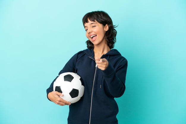 Jeune femme de joueur de football de race mixte isolée sur fond bleu pointant vers l'avant avec une expression heureuse