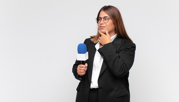 Jeune femme journaliste souriant avec une expression heureuse et confiante avec la main sur le menton, se demandant et regardant sur le côté