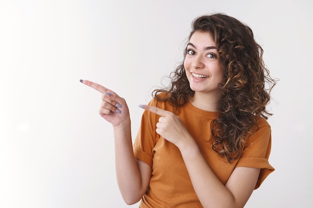 Jeune femme joyeuse et amicale présentant son meilleur ami pointant les doigts vers la gauche tournant joyeusement la caméra souriante suggérant une promotion d'espace vide cool, debout sur fond blanc