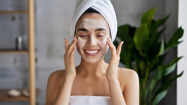 Photo une jeune femme joyeuse applique un gommage naturel sur le visage, touche les joues, garde les yeux fermés, porte une serviette.