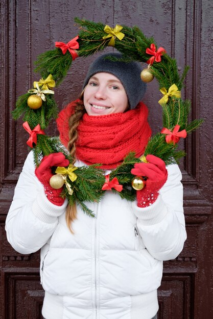 Jeune femme joyeuse attrayante en gants rouges et veste blanche tenant une couronne de Noël devant la porte et souriant