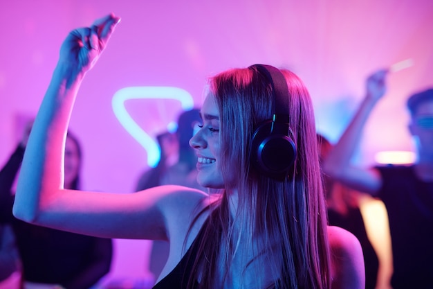 Jeune femme joyeuse aux longs cheveux blonds claquant des doigts tout en écoutant de la musique dans des écouteurs devant une foule de danseurs à la fête