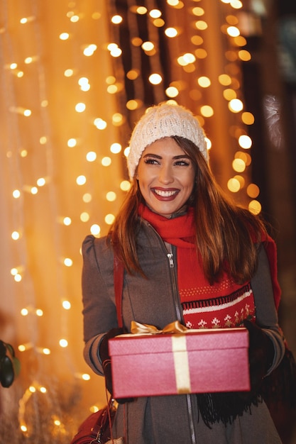 Une jeune femme joyeuse avec une boîte cadeau rouge s'amuse dans la rue de la ville au moment de Noël.