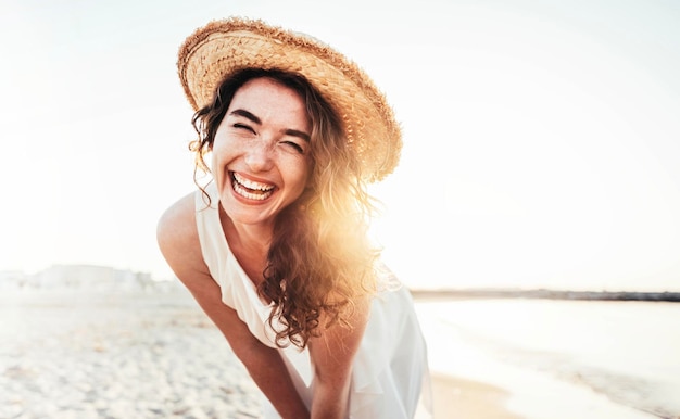 Jeune femme joyeuse en chemise blanche portant un chapeau souriant à la caméra sur la plage
