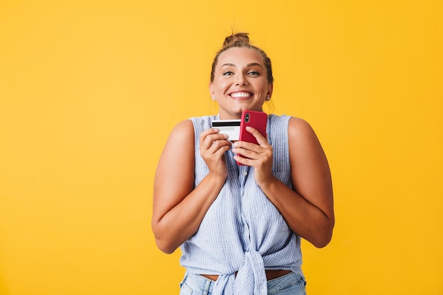 Jeune femme joyeuse en chemise regardant joyeusement à huis clos avec carte de crédit et téléphone portable dans les mains sur fond jaune isolé