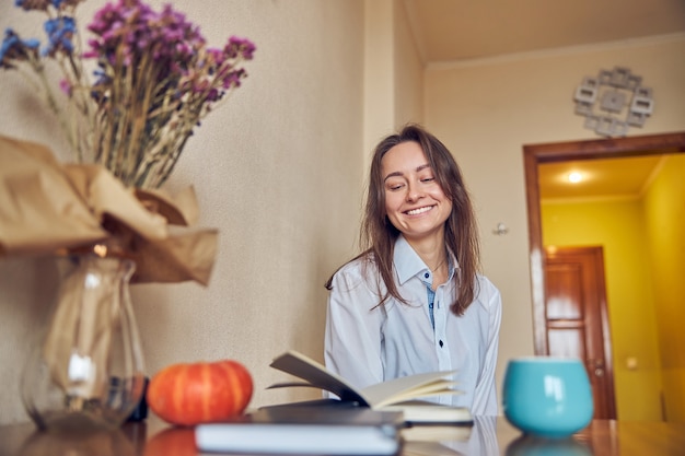 Une jeune femme joyeuse, confiante et heureuse savoure le thé en lisant un livre dans sa cuisine à la maison