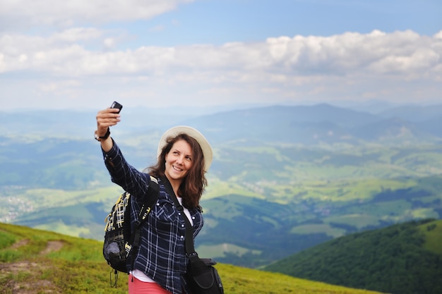 Jeune femme joyeuse dans un chapeau fait selfie au sommet d'une montagne en été