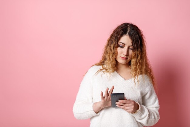 Une jeune femme joyeuse debout isolée sur fond rose avec un smartphone dans les mains