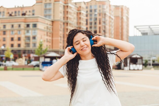 Une jeune femme joyeuse et heureuse avec des dreadlocks vêtue d'un t-shirt blanc dansant en écoutant de la musique avec des écouteurs se reposant dans un parc de la ville marchant le long d'une ruelle Concept de style de vie urbain