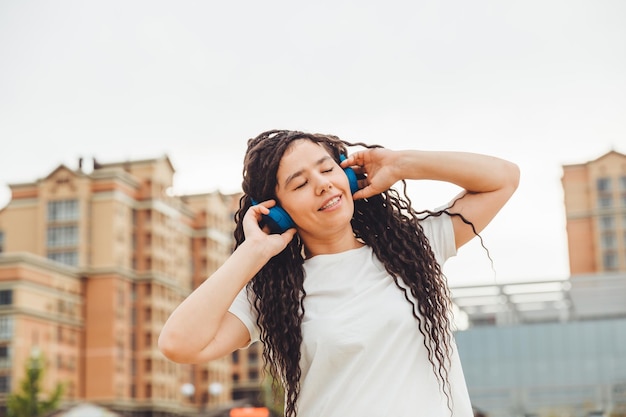 Une jeune femme joyeuse et heureuse avec des dreadlocks vêtue d'un t-shirt blanc dansant en écoutant de la musique avec des écouteurs se reposant dans un parc de la ville marchant le long d'une ruelle Concept de style de vie urbain