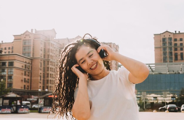 Une jeune femme joyeuse et heureuse avec des dreadlocks vêtue d'un t-shirt blanc dansant en écoutant de la musique avec des écouteurs se reposant dans un parc de la ville marchant le long d'une ruelle Concept de style de vie urbain