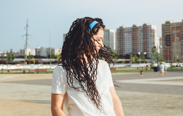 Une jeune femme joyeuse et heureuse avec des dreadlocks vêtue d'un t-shirt blanc dansant en écoutant de la musique avec des écouteurs se reposant dans un parc de la ville marchant le long d'une ruelle Concept de style de vie urbain