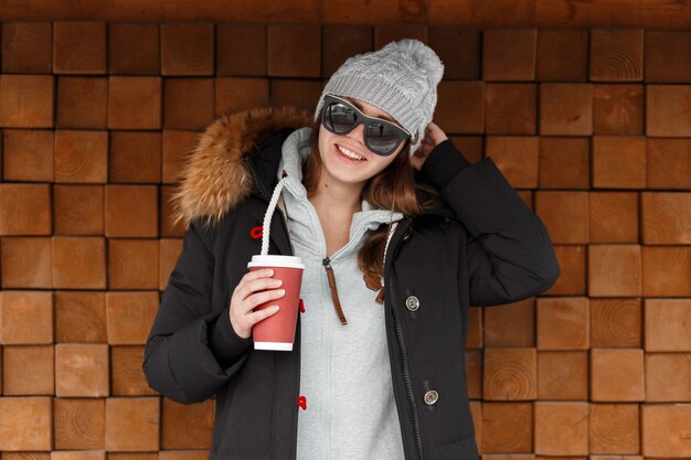 Jeune femme joyeuse hipster dans un chapeau tricoté gris à lunettes de soleil dans une veste élégante est debout et tenant une tasse rouge avec une boisson chaude près d'un mur en bois vintage. Funny girl passe le week-end.