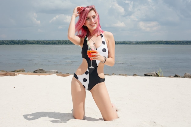 Jeune femme joyeuse en maillot de bain, grillant avec un cocktail, buvant une boisson fraîche sur la plage, au bord de la mer. Notion de vacances.