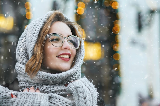 Une jeune femme joyeuse porte une casquette grise et des lunettes marchant dans la ville pendant les chutes de neige. Espace libre