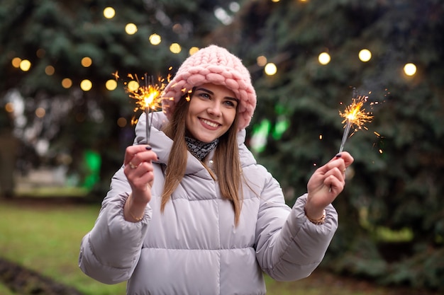 Une jeune femme joyeuse porte un chapeau et un manteau tricotés roses s'amusant avec des cierges magiques dans la rue près de l'arbre de Noël