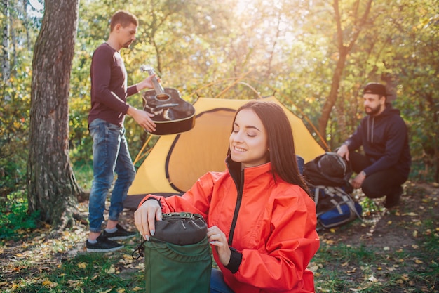 Jeune femme joyeuse et positive sourit et détient le sac à dos. Elle le regarde. Premier jeune homme tenir une guitare noire. Un autre assis près de la tente et du sac à dos.