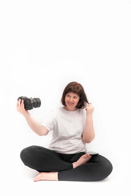 Une jeune femme joyeuse pratique le yoga et se prend en photo avec un appareil photo. Portrait de jeune fille en demi-lotus pose sur fond blanc.