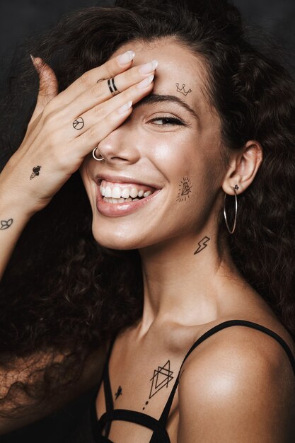 jeune femme joyeuse avec un tatouage souriant et couvrant son œil isolé sur un mur noir