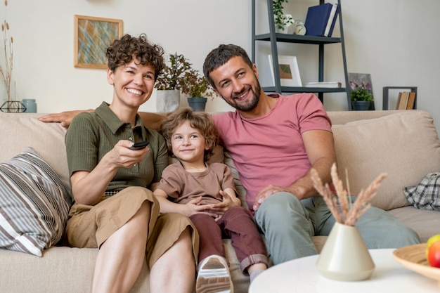 Jeune femme joyeuse avec télécommande, son mari et leur mignon petit fils assis sur un canapé confortable en face de la télévision à la maison