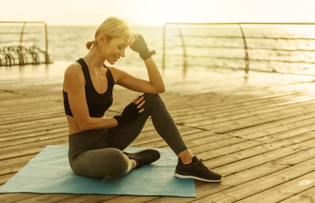 Jeune femme joyeuse en tenue de sport est assise sur un tapis sur la plage au lever du soleil. Entraînement du matin